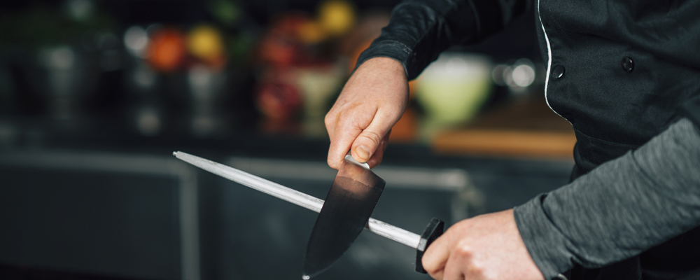 Male chef in black uniform sharpening the knives in the kitchen.